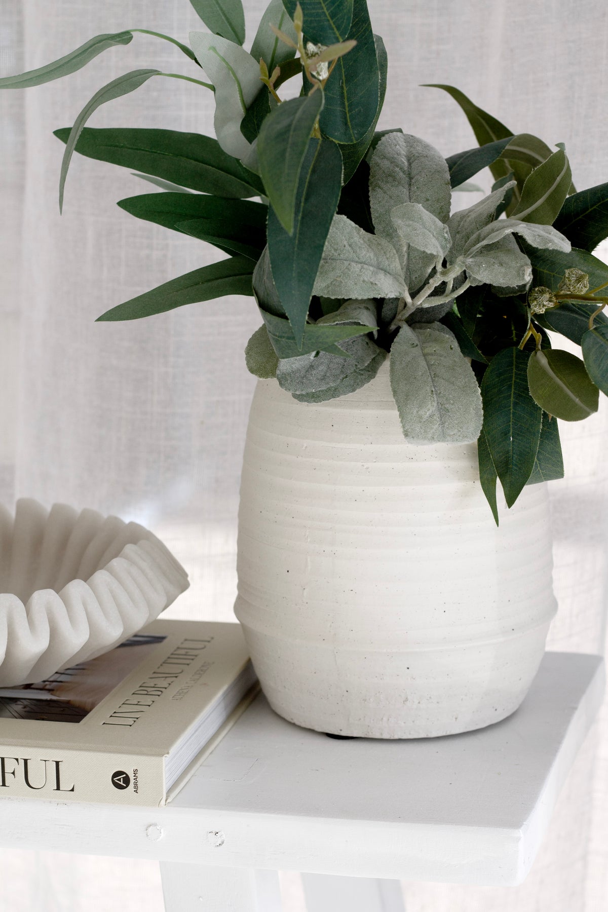 Alma vase in chalk holding leafy green stems beside a ruffled white bowl on a coffee table book