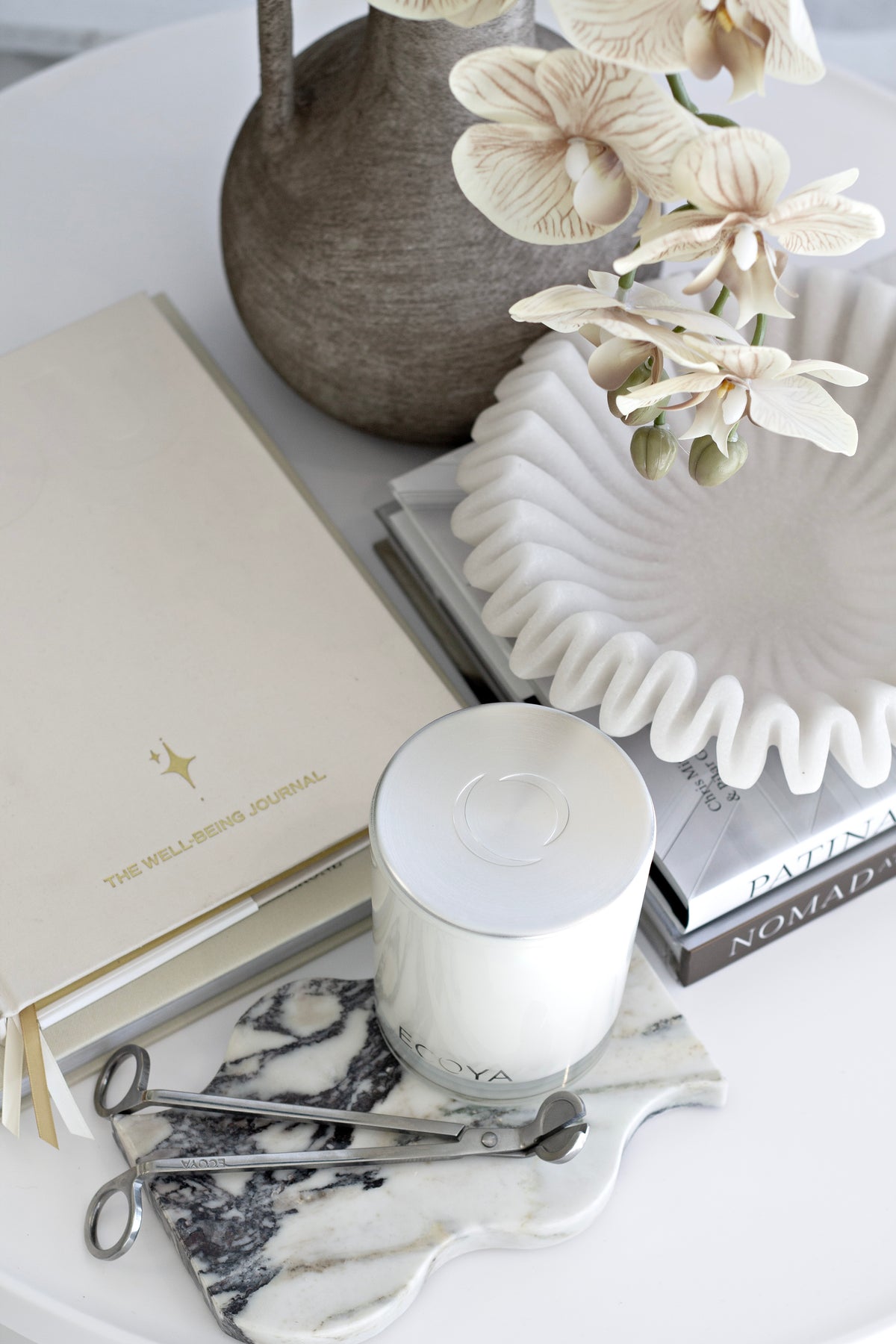 Orchids in a dark rustic vase with a fluted dish, a candle and silver scissors on books