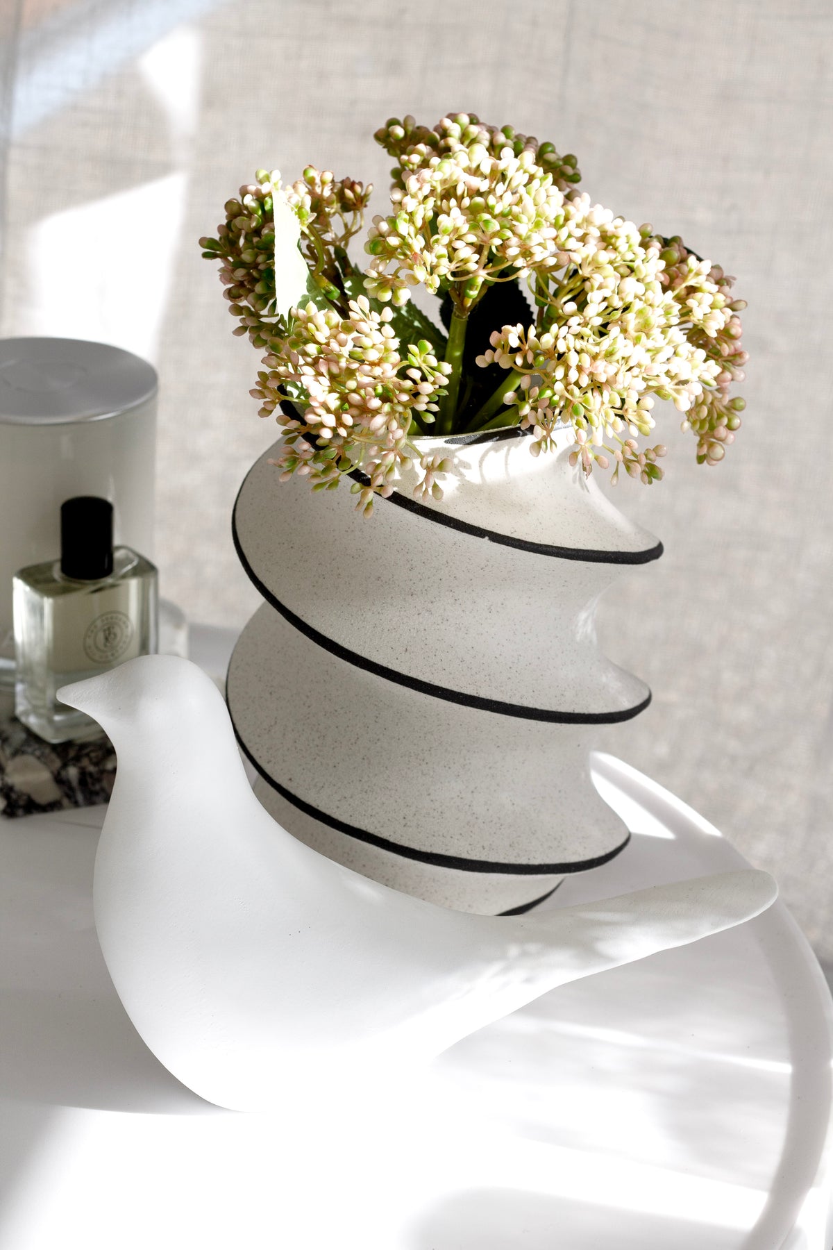White bird figurine in sunlight beside a striped speckled vase of white blossom and a perfume bottle