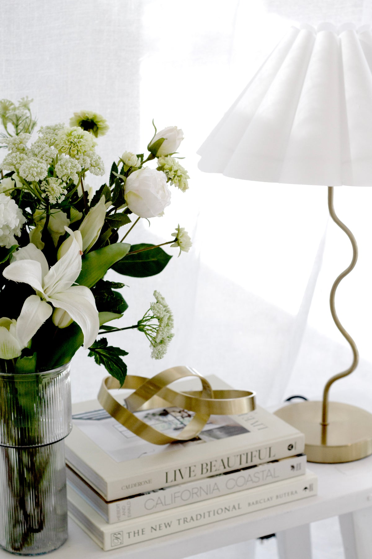White lilies and dill ammi in a glass vase beside books and a wavy lamp