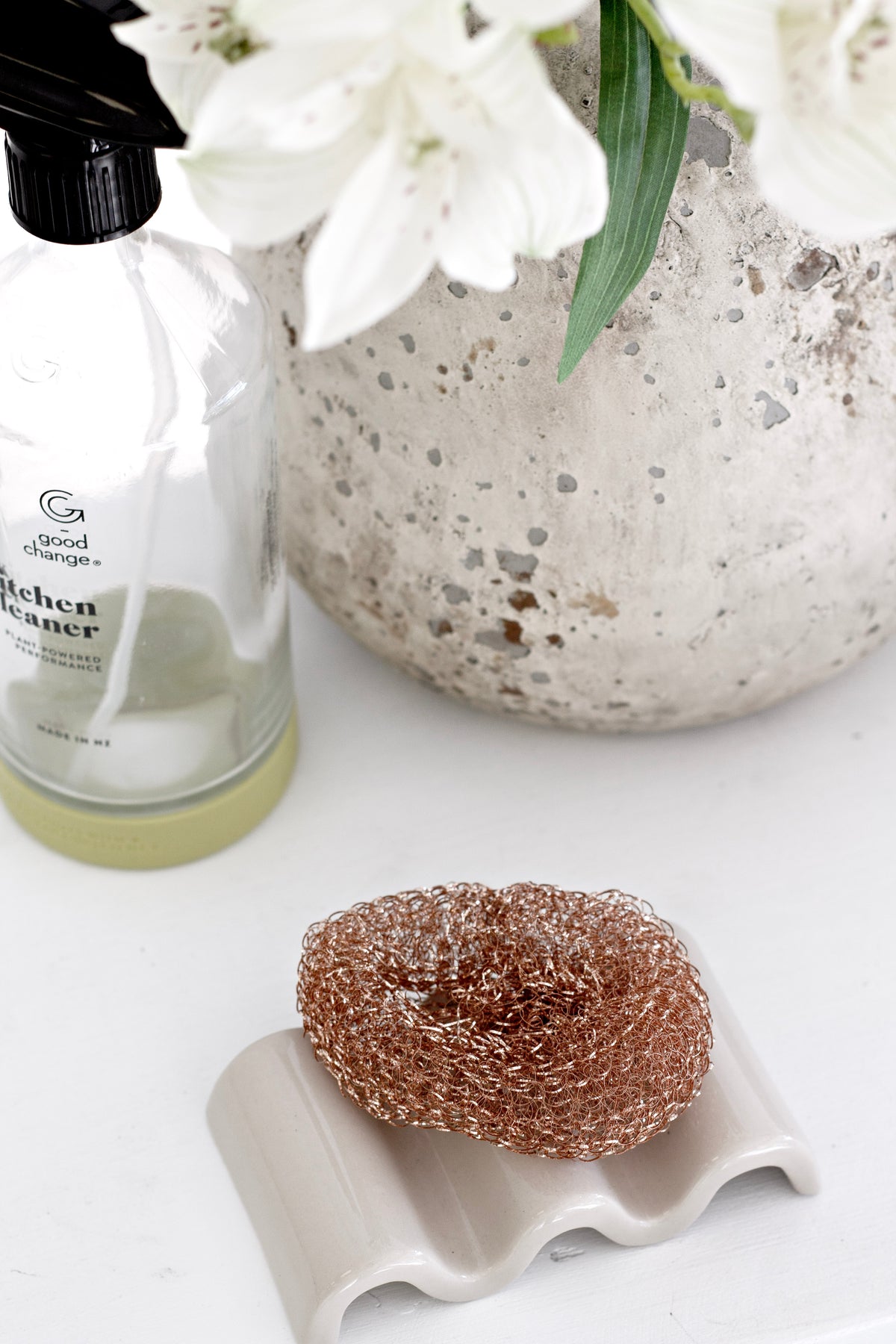 Copper sponge on a white wavy soap dish beside a glass bottle and white flowers
