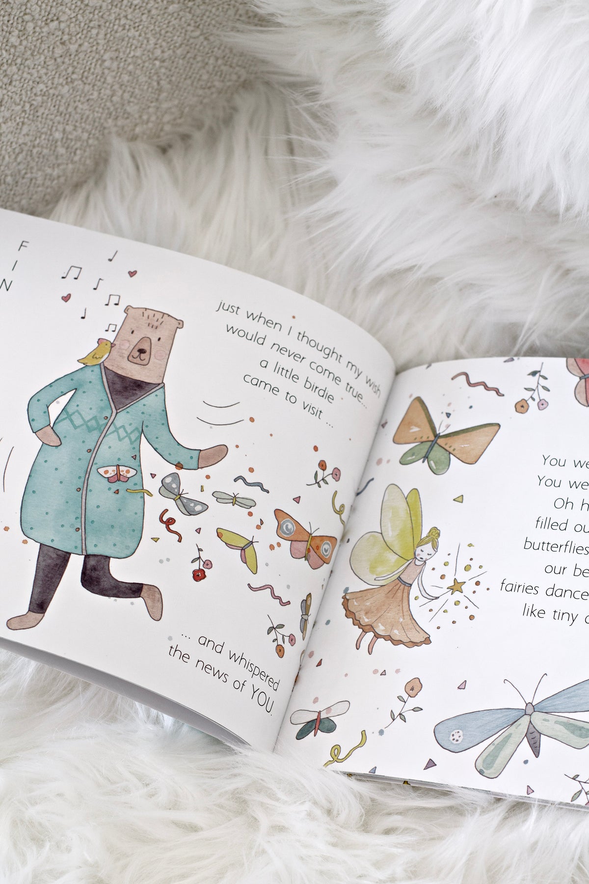 Before You Were Here open to a watercolour spread of a bear in a coat and butterflies, on a white rug