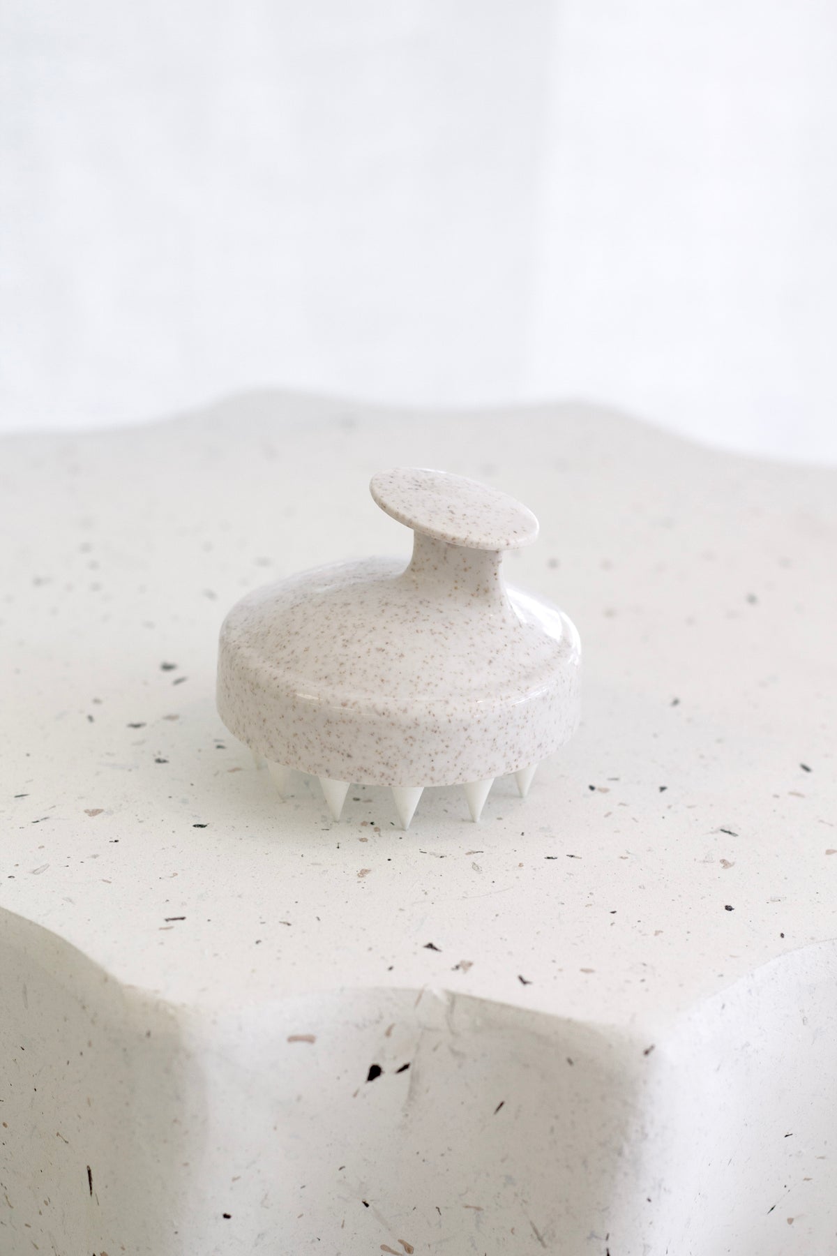 Speckled cream scalp massager standing bristles down on a textured white plaster surface