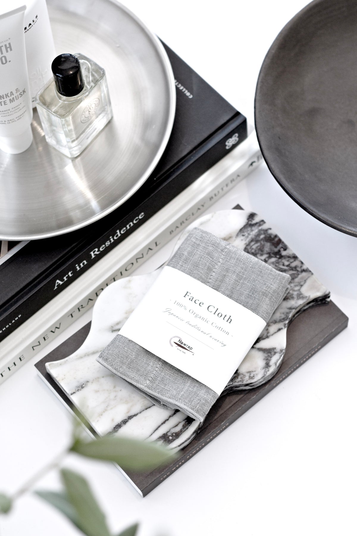 Charcoal face cloth in its band on a stack of books beside a perfume bottle and a metal tray