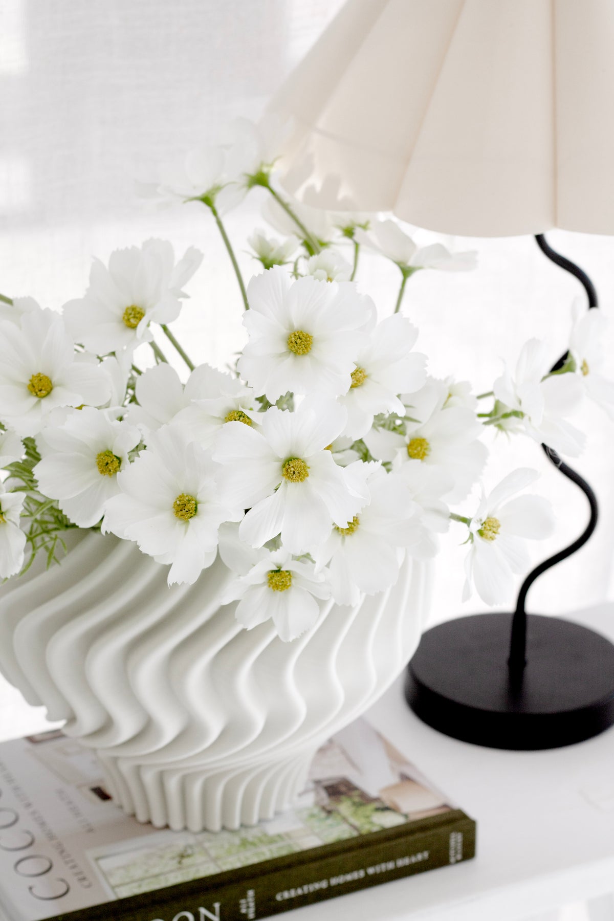 Bunch of white cosmos in a white wavy vase on stacked books under a pleated lamp