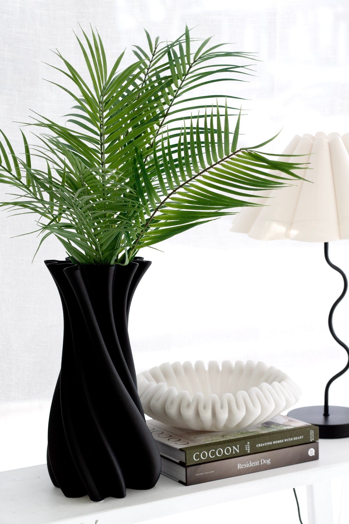 Black Camila vase with palm fronds beside a table lamp and a stack of books