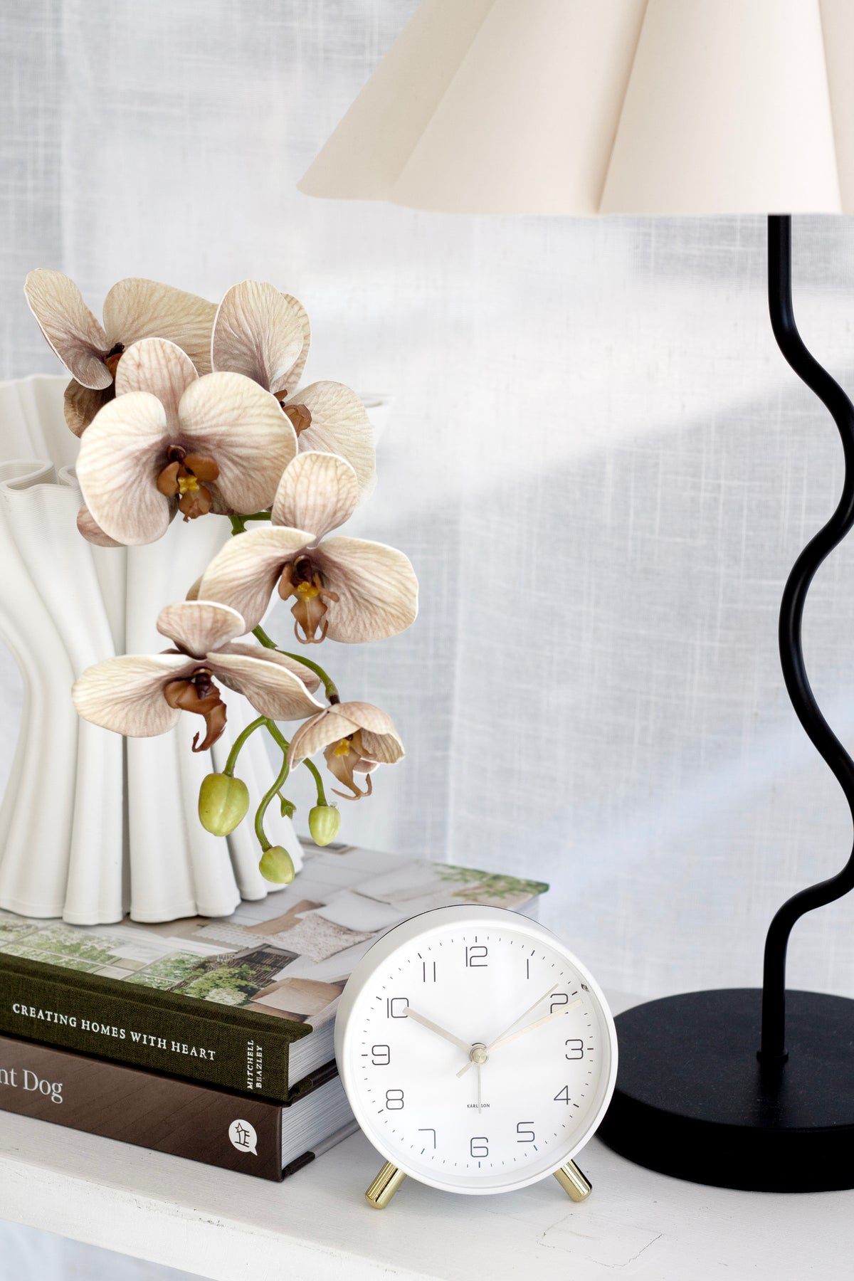White Karlsson Lofty alarm clock on books beside dried flowers and a black lamp