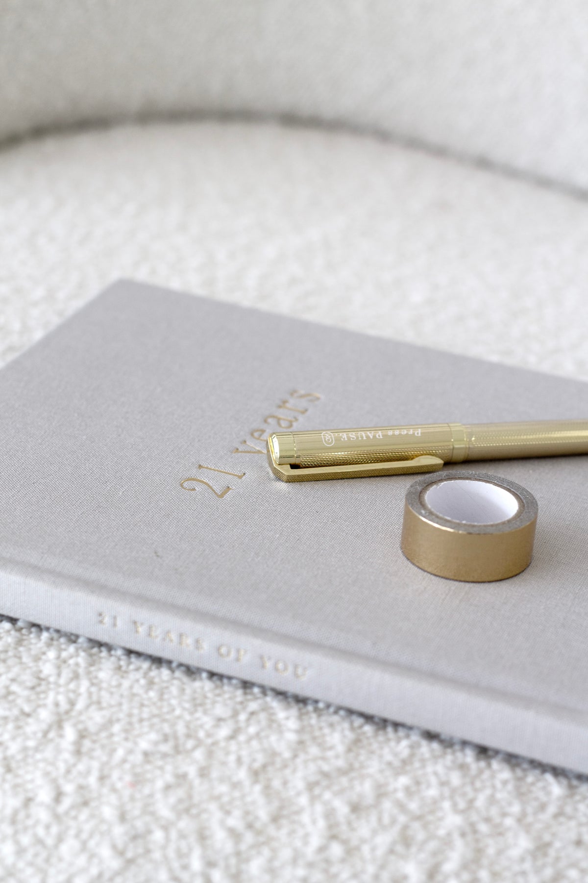 Grey 21 Years journal with a gold pen and a brass object on a boucle surface