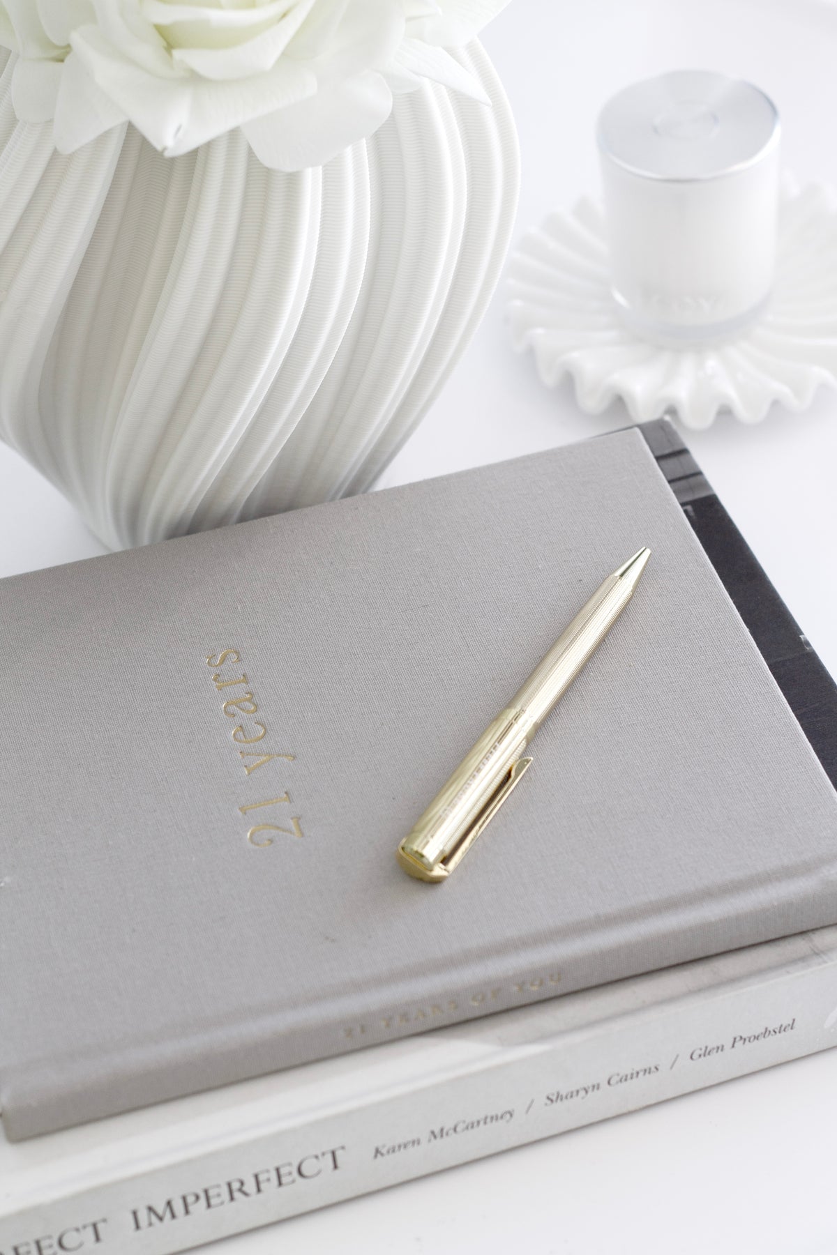 Grey 21 Years journal with a gold pen on a white table beside a sculptural vase