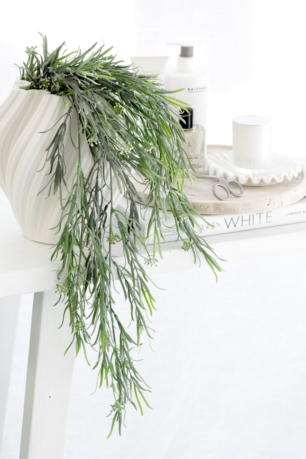 White Amelie fluted vase with trailing green foliage on a white console beside skincare