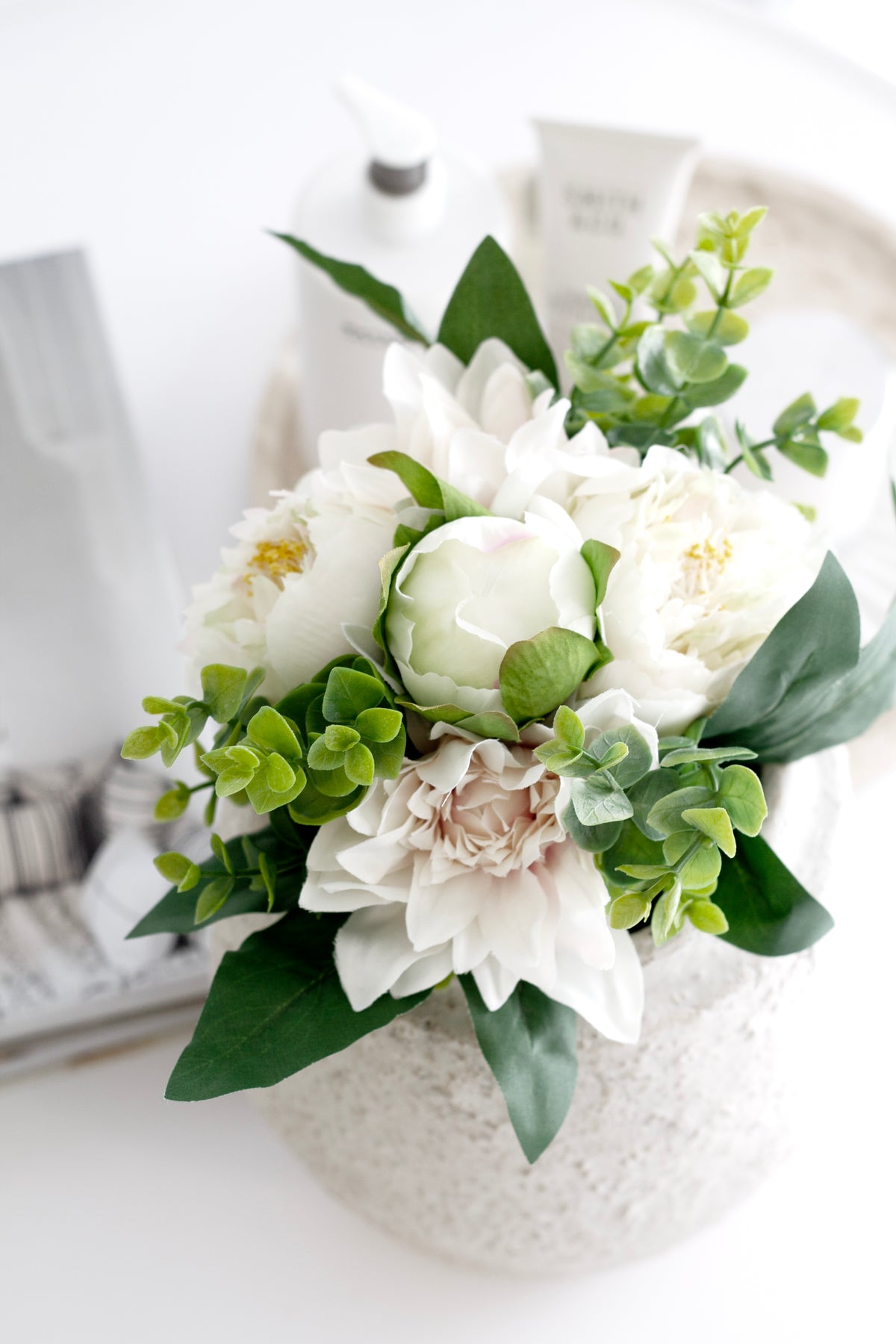 Blush faux dahlia and peony bouquet in a speckled white vase with bottles behind