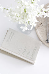 Book titled 'All That You Deserve' with white flowers on a light surface