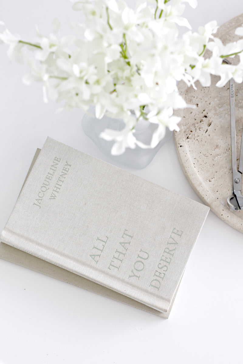 Book titled 'All That You Deserve' with white flowers on a light surface