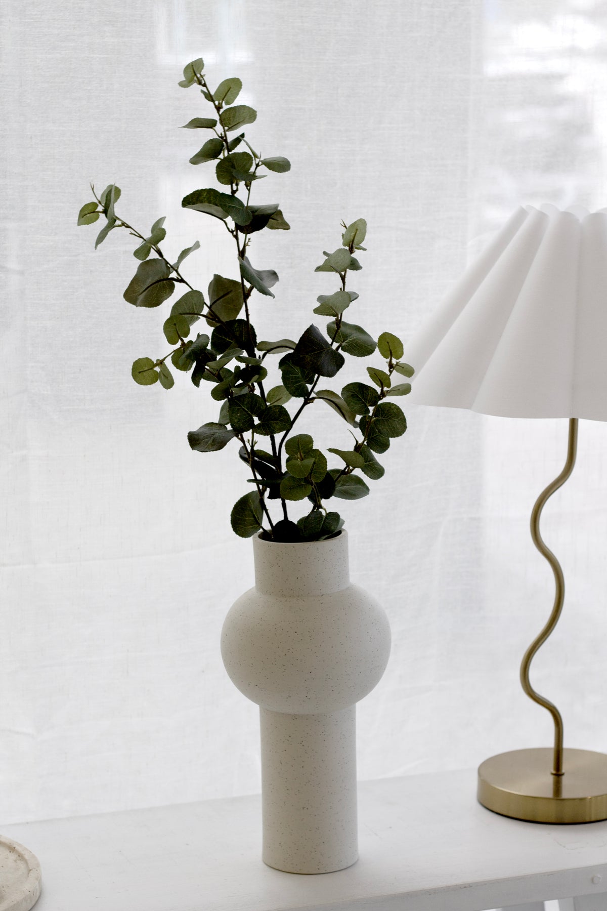 Faux eucalyptus in a sculptural white vase beside a pleated lamp