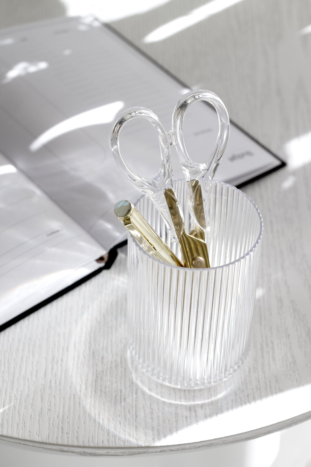 Clear fluted tumbler used as a desk tidy holding gold scissors and pens in afternoon sun