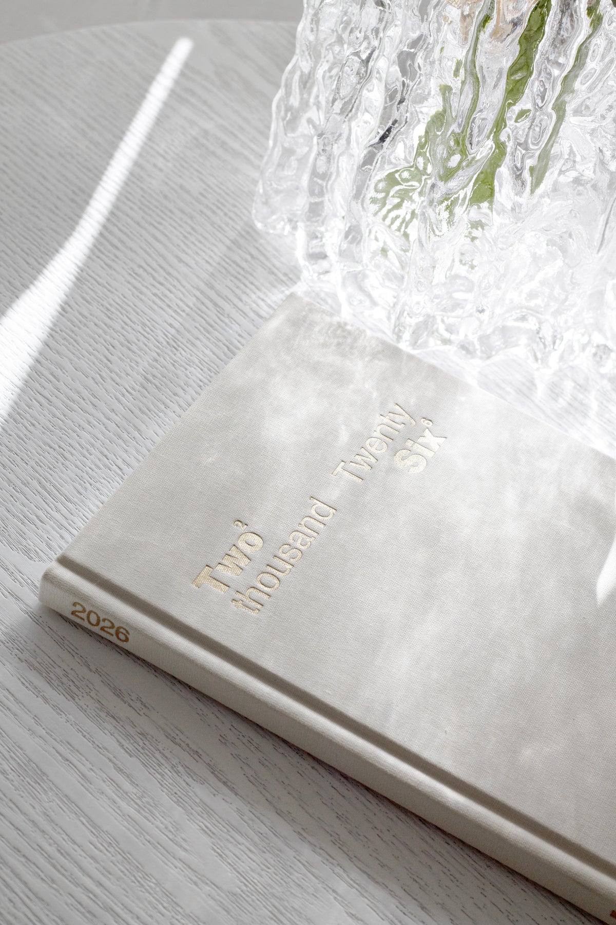 Clear Glacier vase of white blooms on a book on a timber surface, backlit by a window