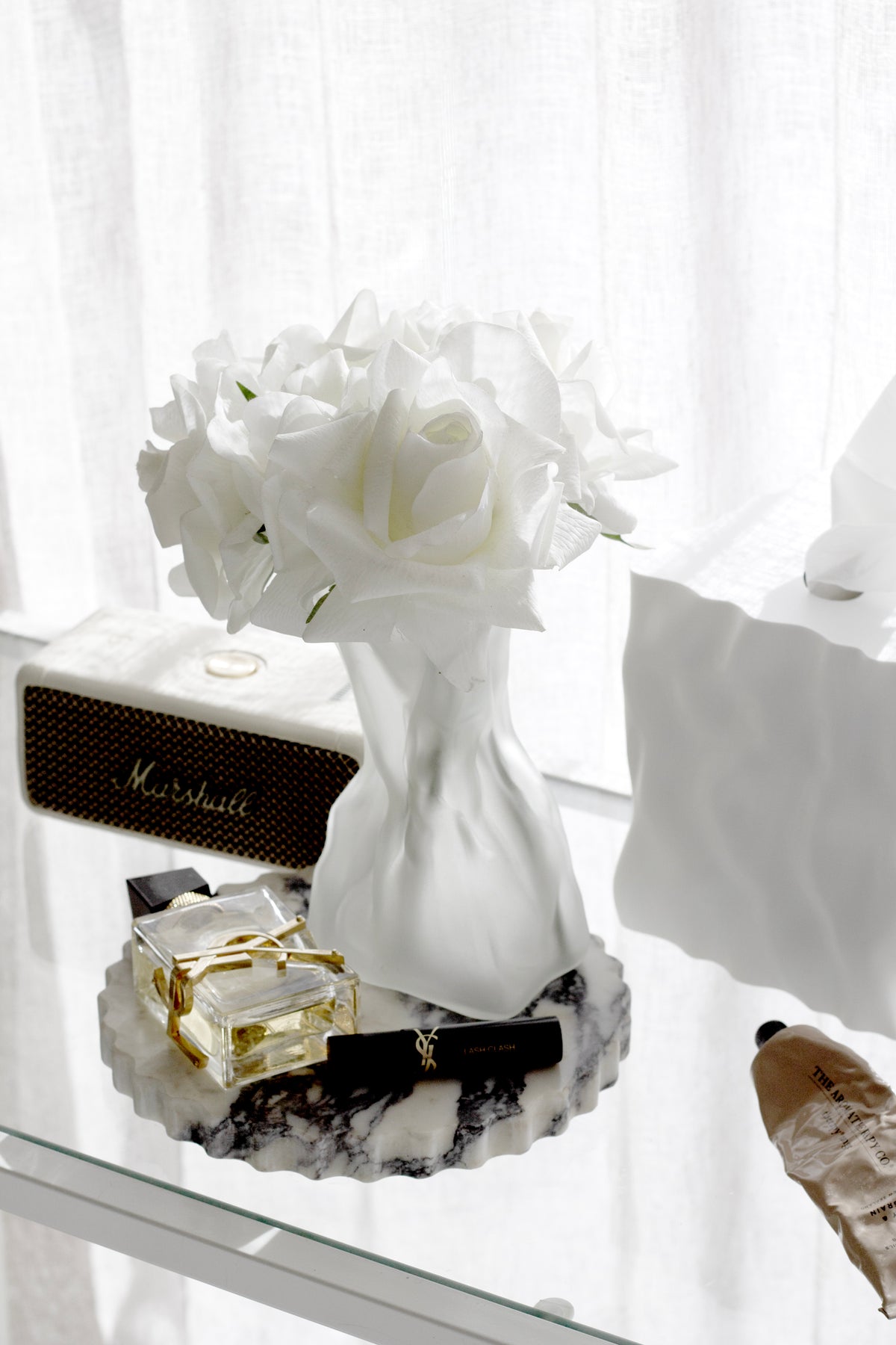 Cream crinkle vase of white roses on a glass shelf beside a jewellery box and gold pieces