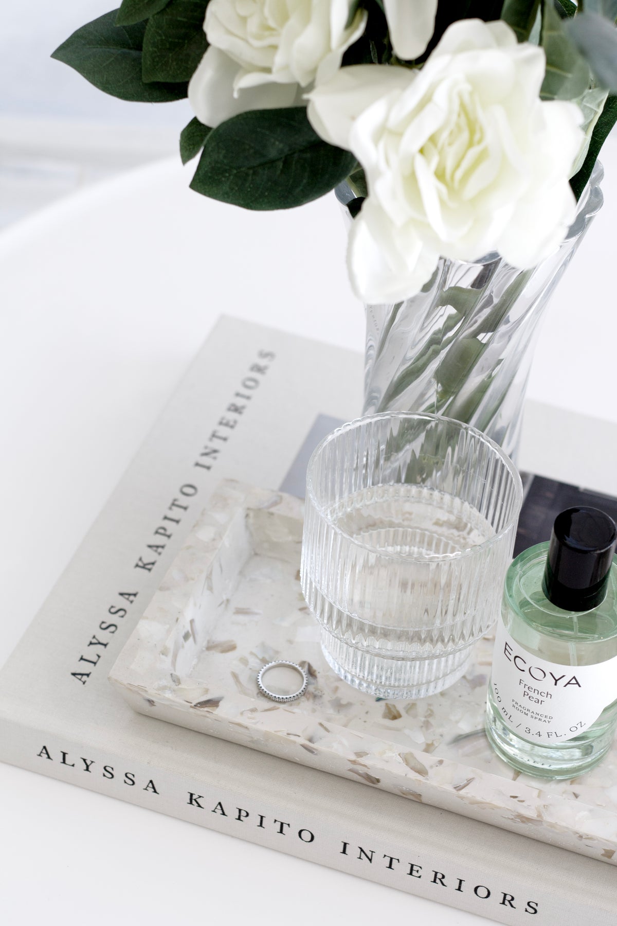 White gardenia in a clear glass vase on a stack of books with a travertine tray and fragrance