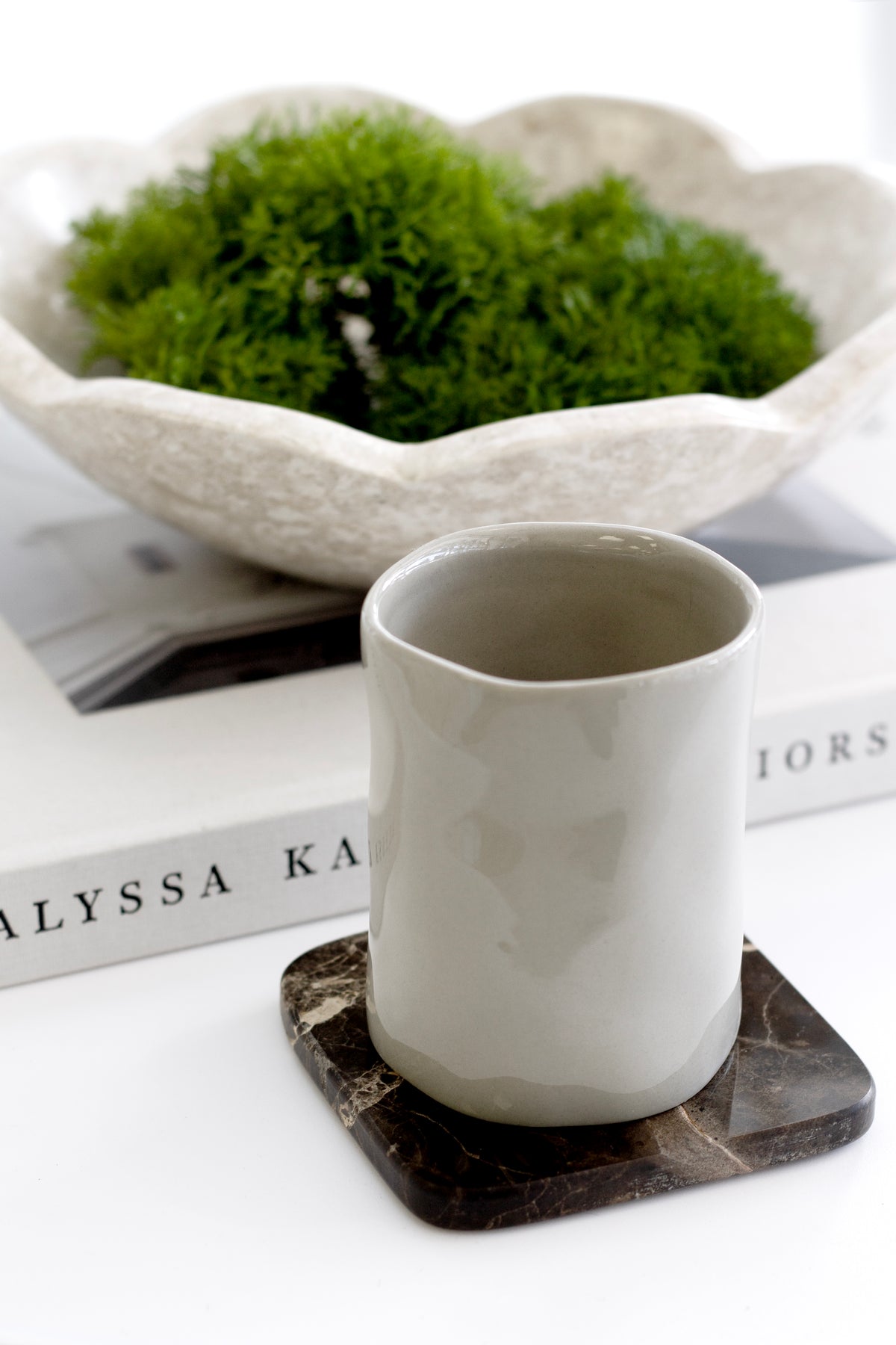 White ceramic tumbler on a dark marble coaster beside a bowl of moss on books