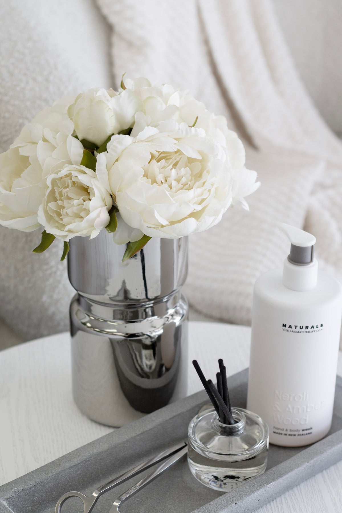 White peonies in the silver vase beside a pump bottle on a grey tray
