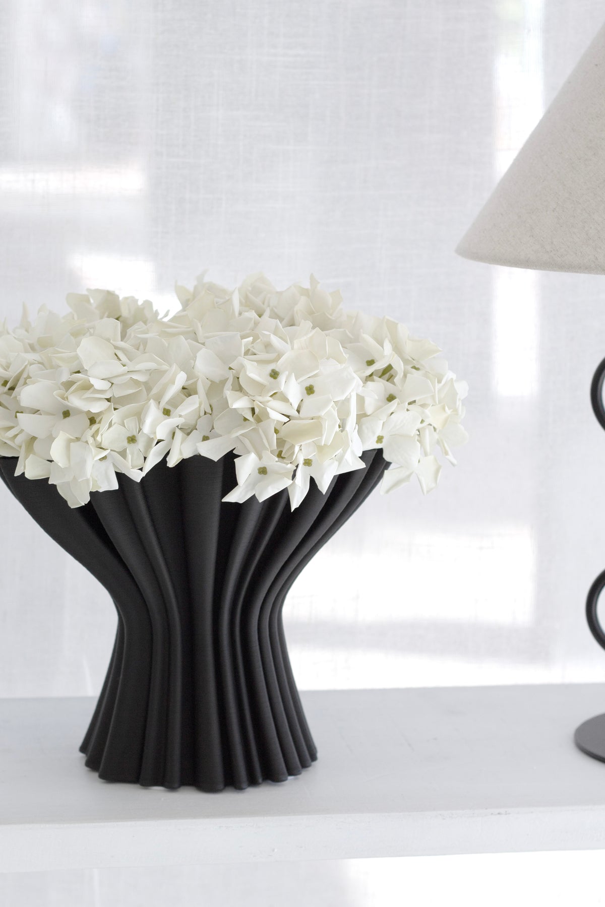 Black Cleo compote vase holding white hydrangeas on a white surface beside a table lamp