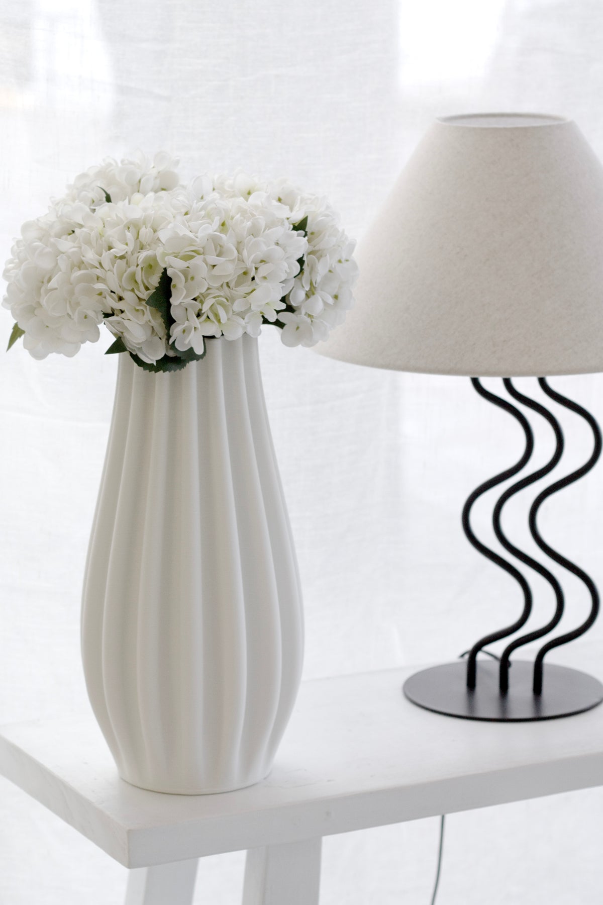 Hydrangeas in a tall fluted vase on a white side table beside a wavy based lamp