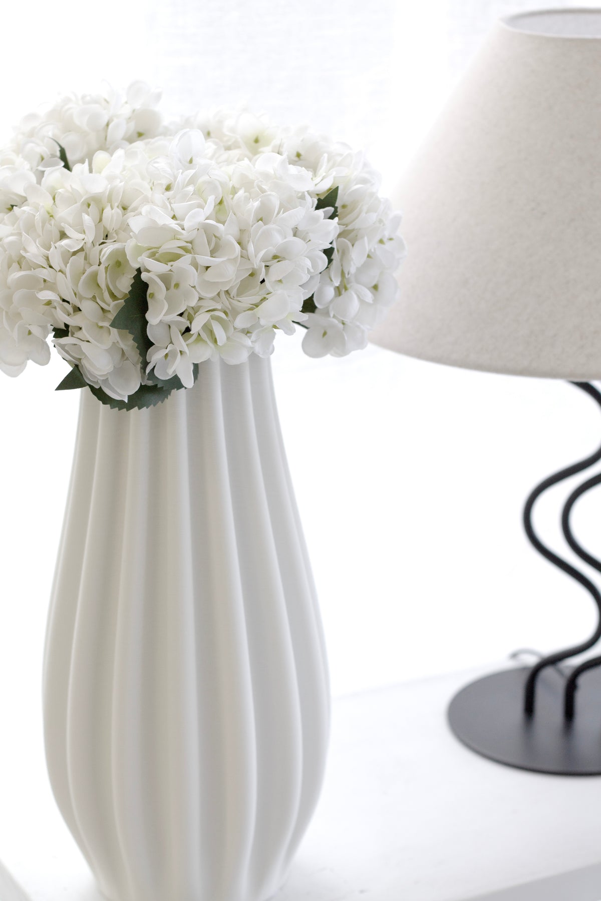 Hydrangeas in a fluted white vase beside a lamp with a black wavy stem