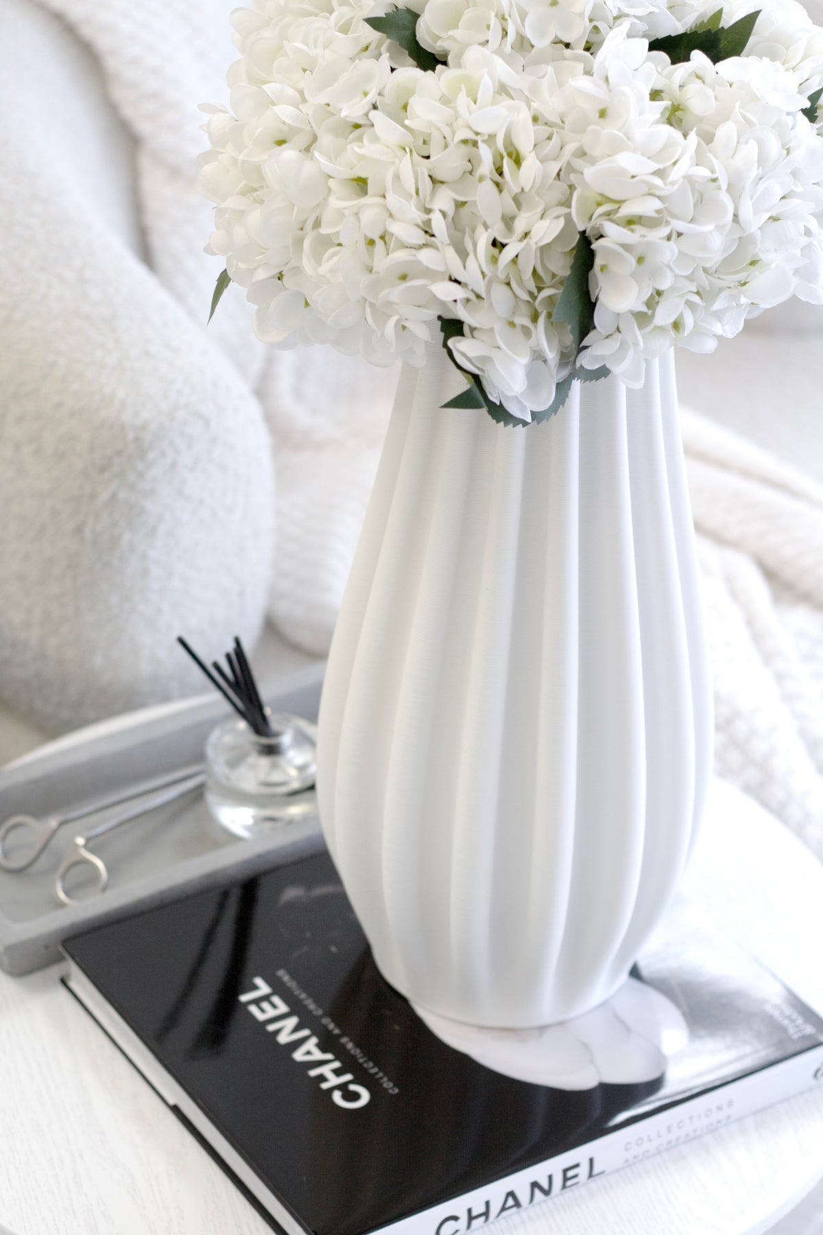 White hydrangeas in a tall ribbed vase on a black book beside a small reed diffuser
