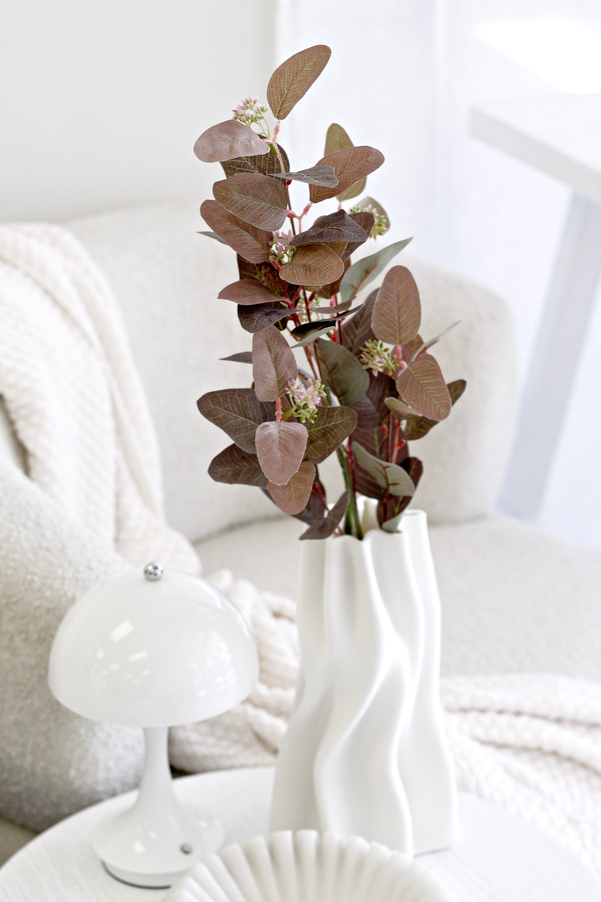 Seeded gum in a sculptural white vase beside a soft throw and a white ornament