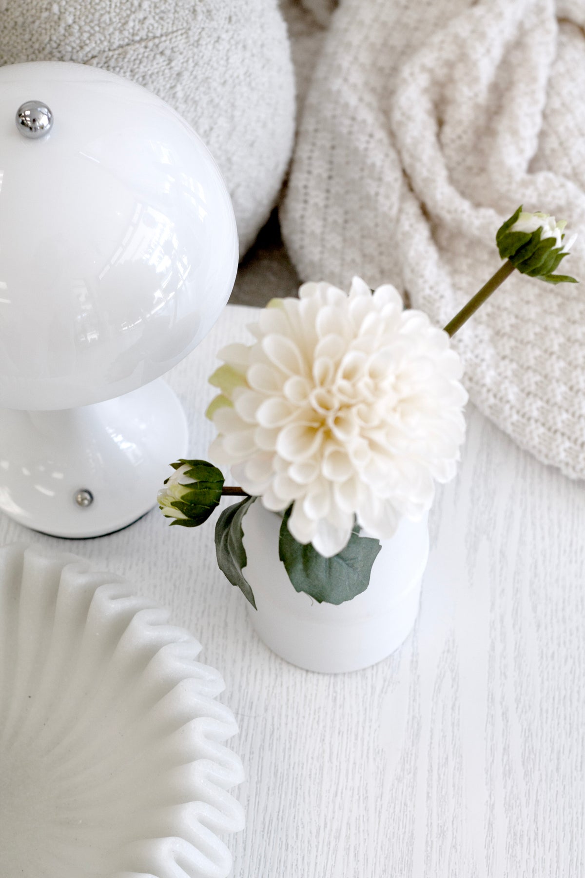 A dahlia in a small white vase beside a round lamp and a fluted dish on pale timber