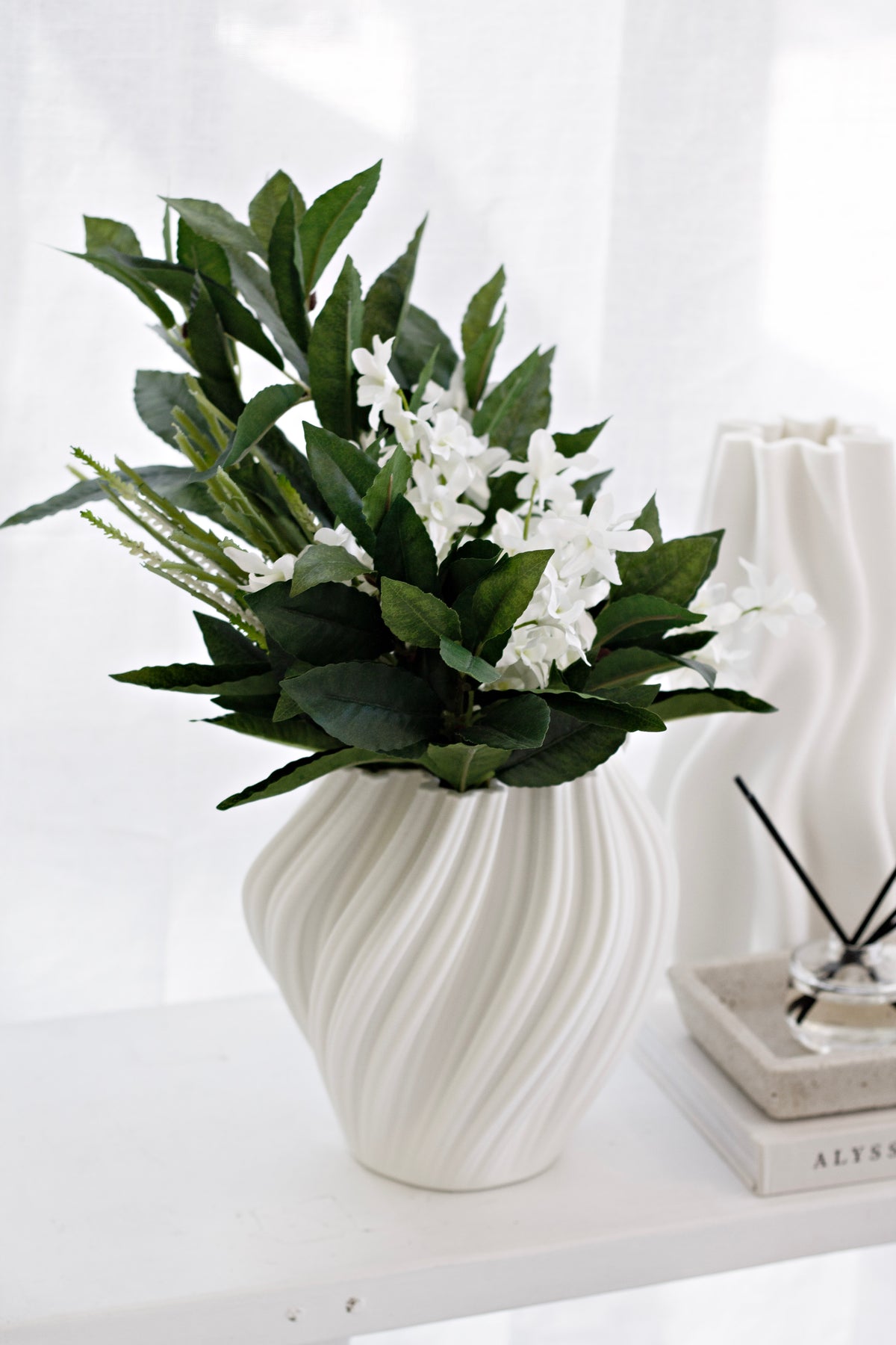 Leafy stems with white flowers in a swirled white vase beside a reed diffuser