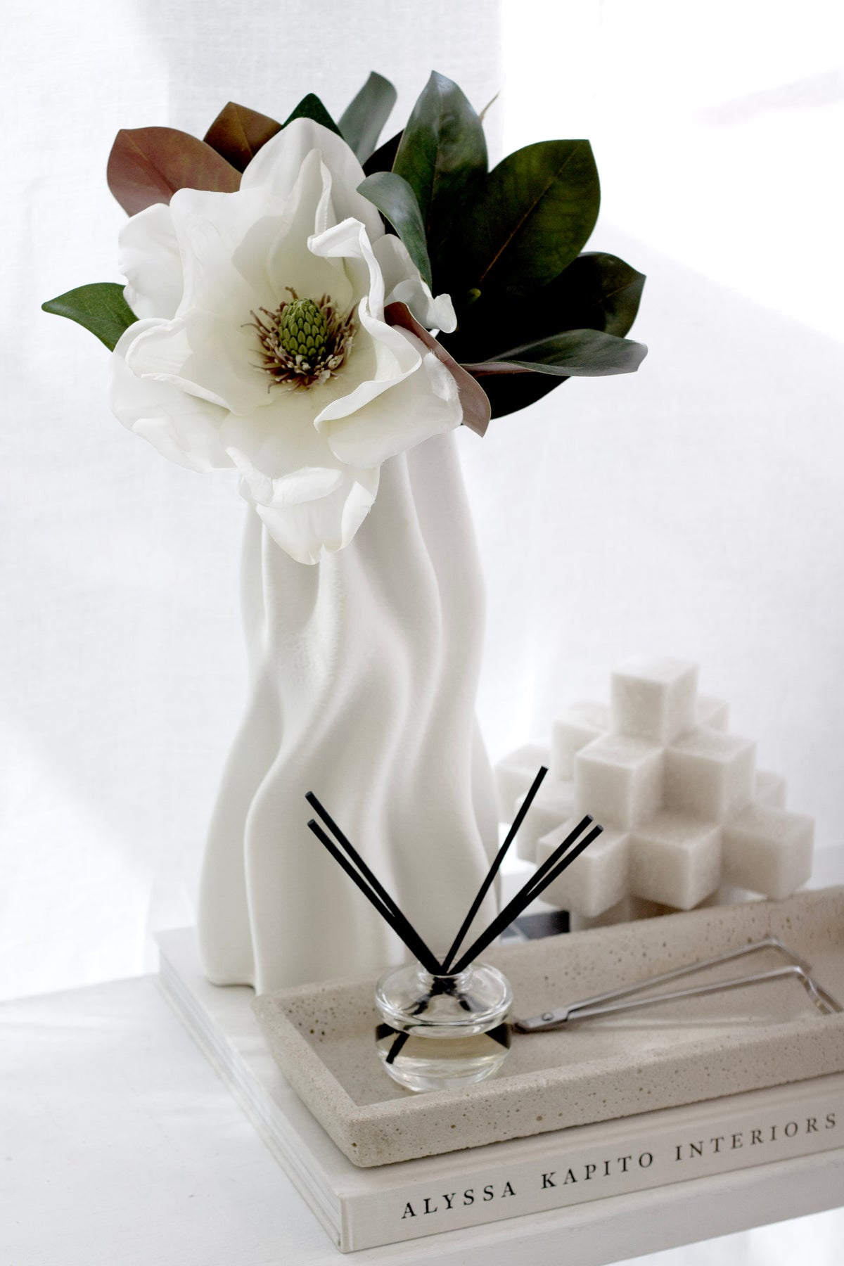 Magnolia leaves with a white magnolia bloom in a wavy vase beside a diffuser