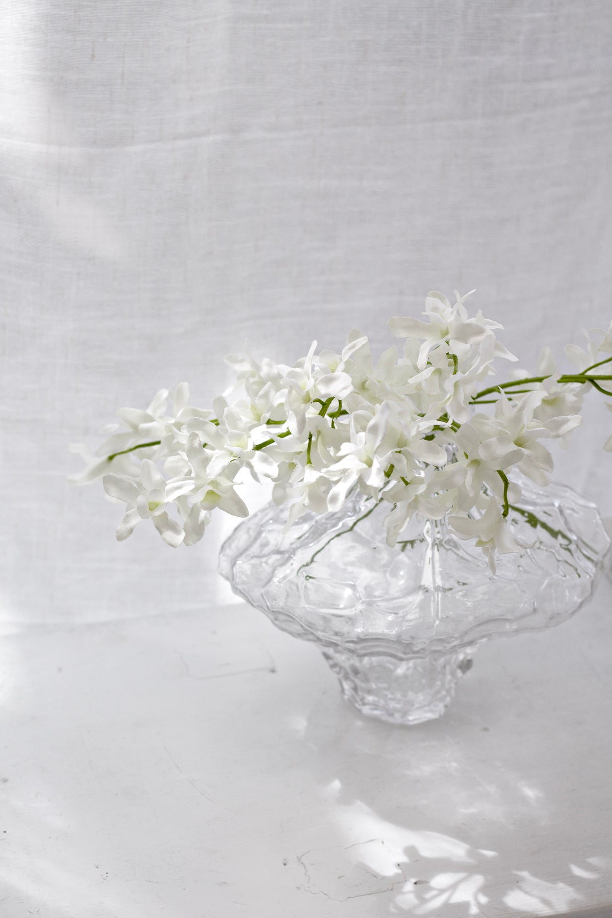 Faux orchid bundle in a footed glass bowl by sheer curtains