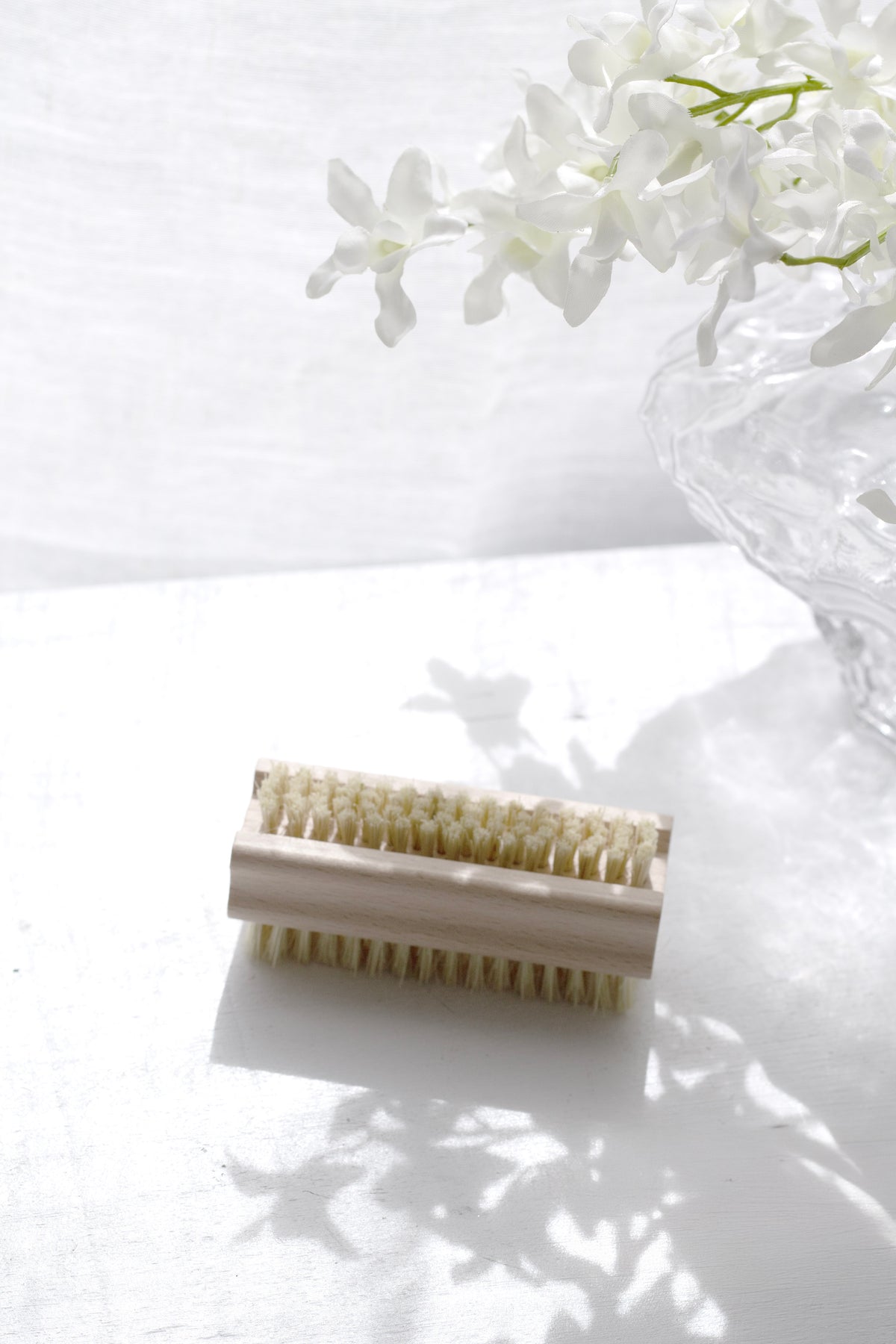 Beechwood nail brush on a white surface in dappled light beside white blossom