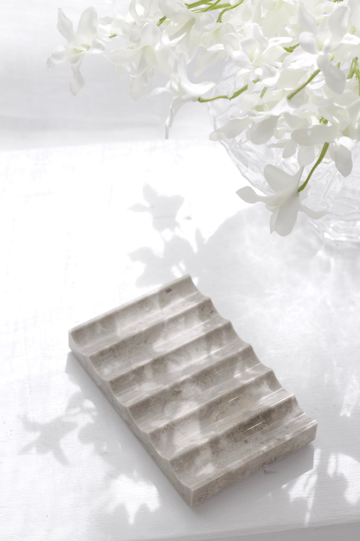 Fluted travertine tray in dappled light on a white surface, white blossom in a glass vase behind