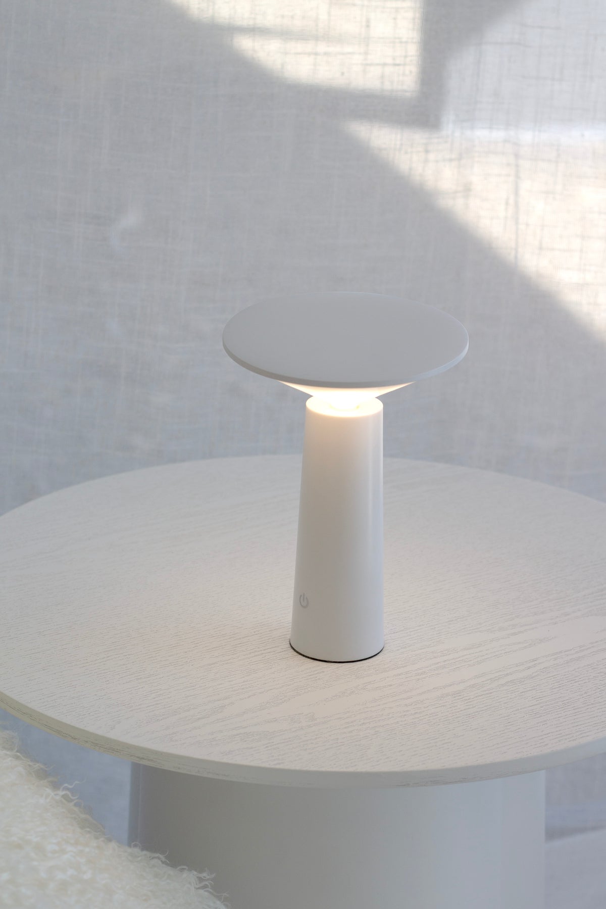 White LED Tilt lamp lit on a pale round table against a sheer curtain