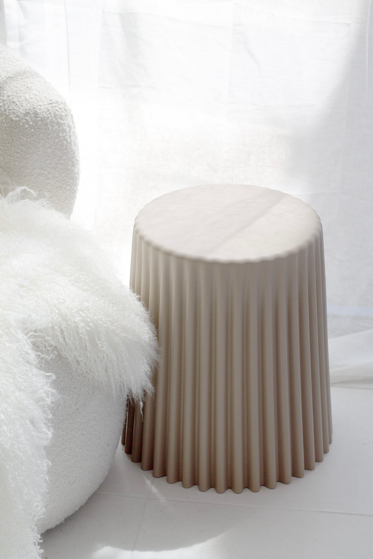 Nude ribbed stool beside a white boucle chair with a sheepskin cushion on a pale floor