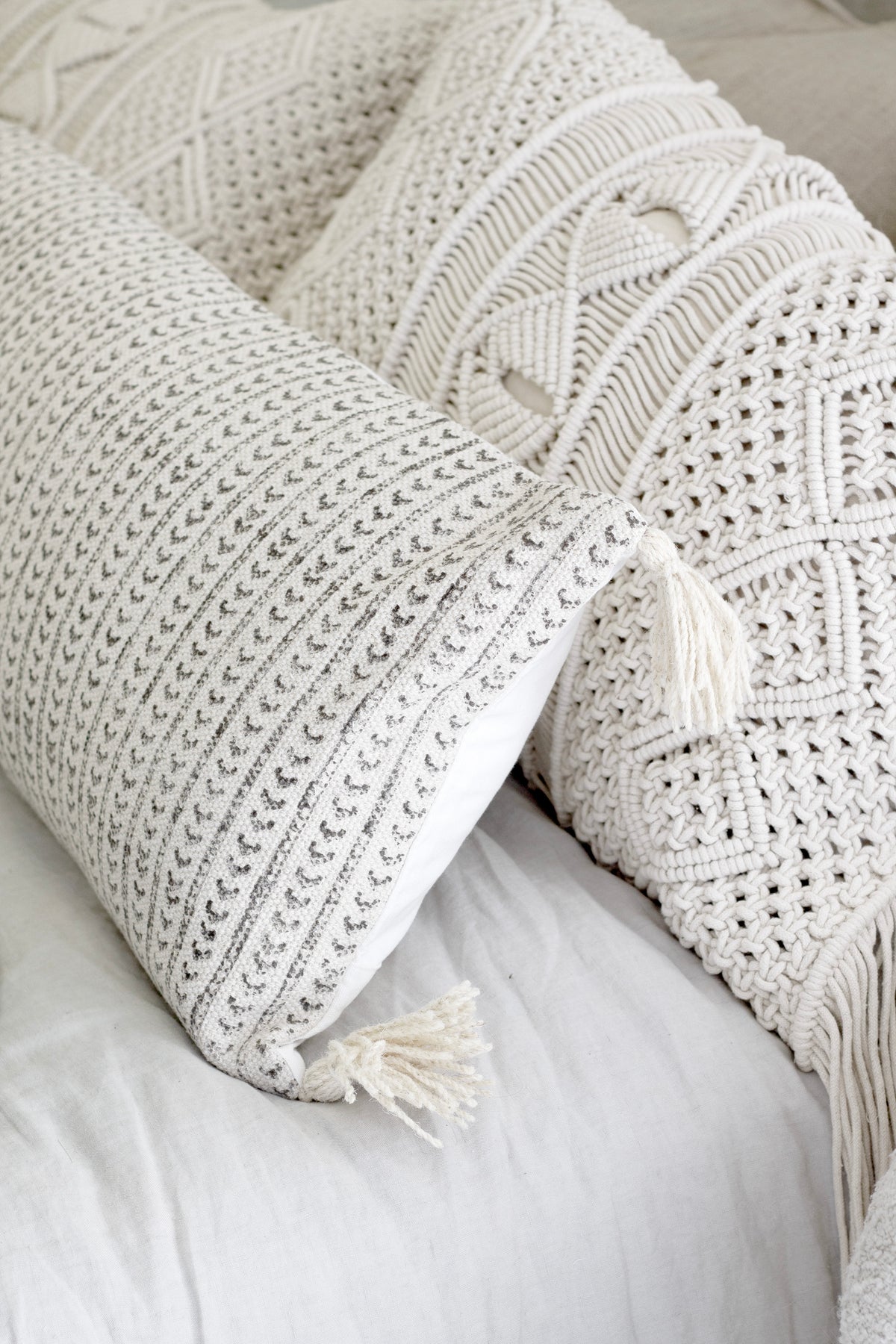 Two macrame cushions layered on a bed, tasselled corners hanging over white linen
