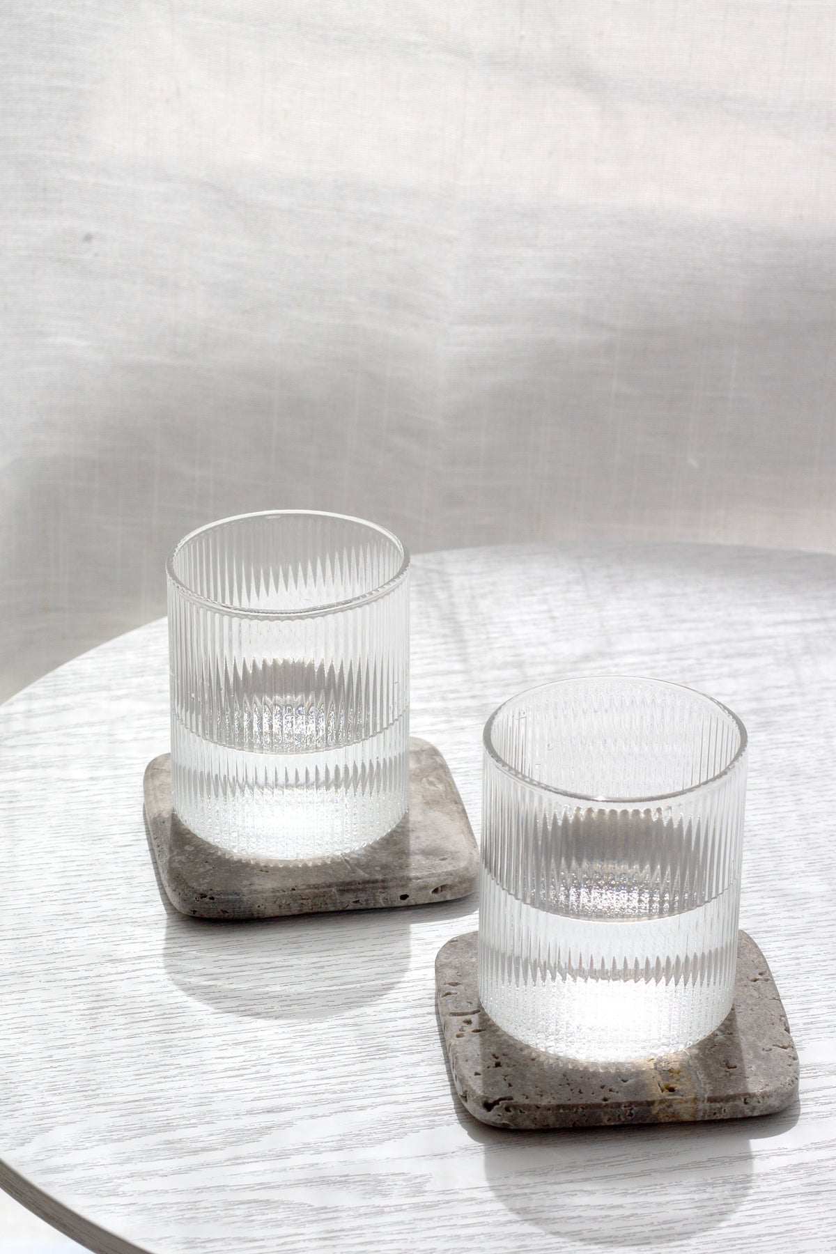 Two ribbed glasses of water on travertine coasters on a white round table