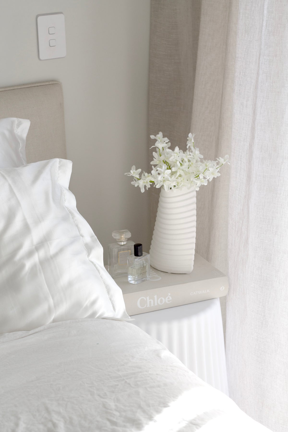 Nude ribbed stool beside an upholstered headboard, holding a vase of white flowers and glass bottles