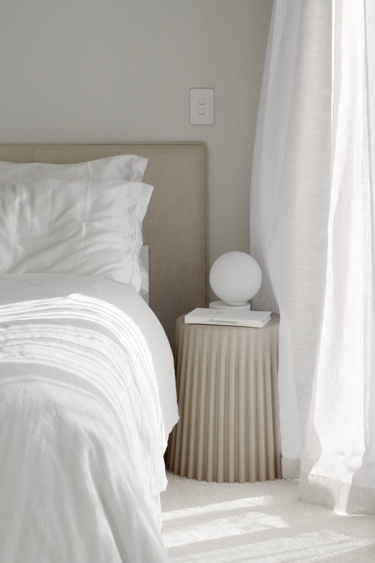 Nude ribbed stool beside a bed in layered white linen, a globe lamp on top in bright sun