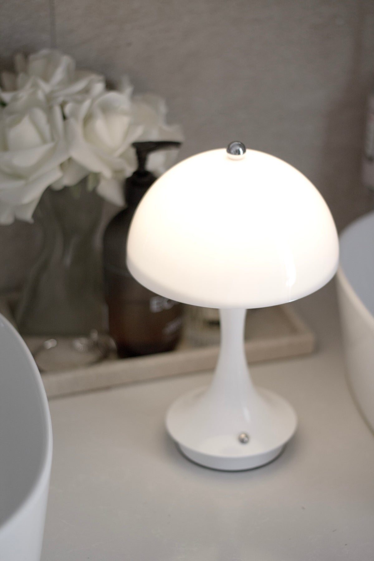 White mushroom lamp lit on a dressing table beside a vase of white roses and a mirror