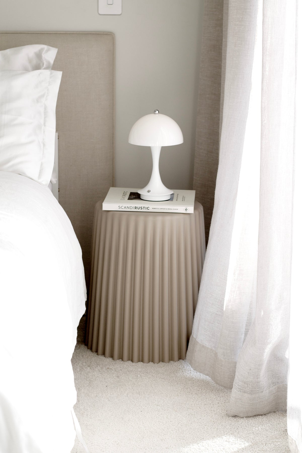 White mushroom lamp lit on a fluted bedside table beside a bed and a sheer curtain