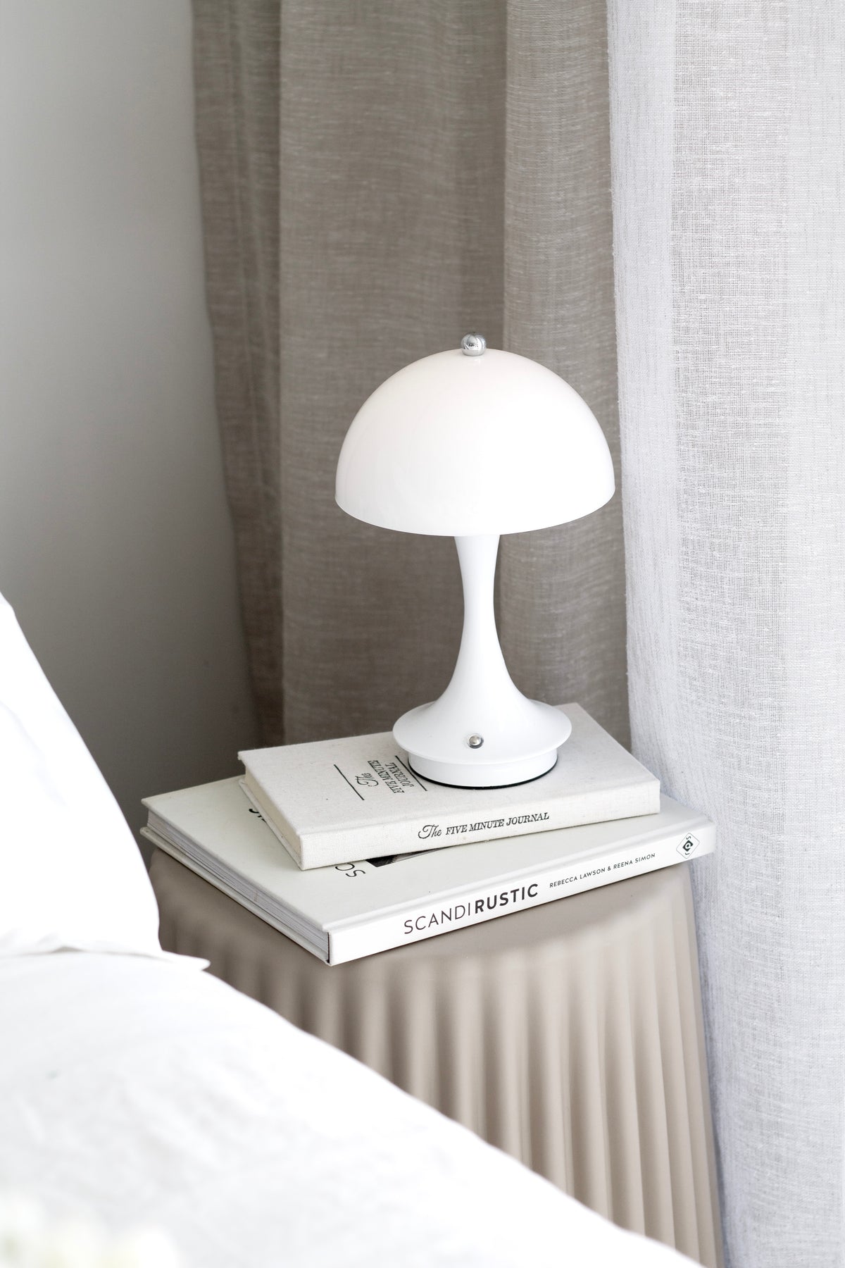 Nude ribbed stool as a bedside table with a mushroom lamp on a stack of books, sheer curtain behind
