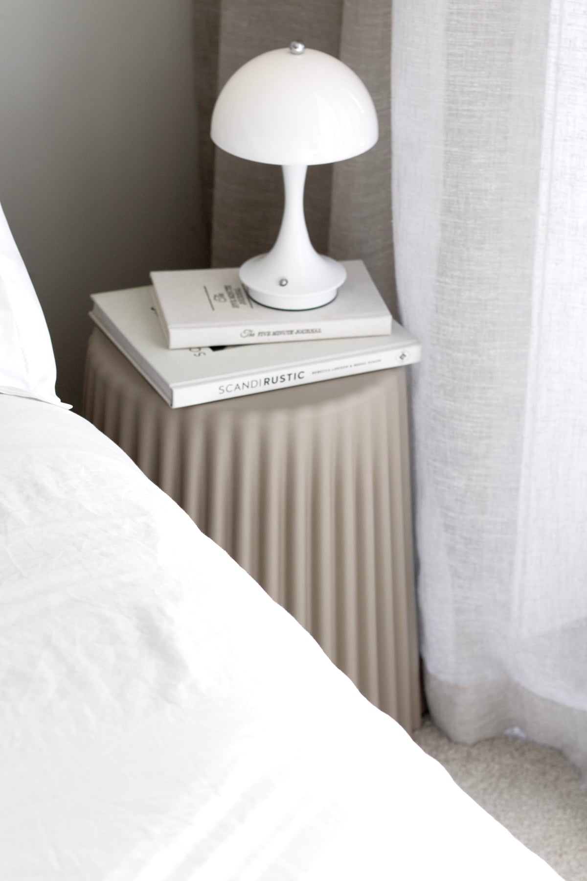 White mushroom lamp unlit on books on a fluted bedside table beside white bedding