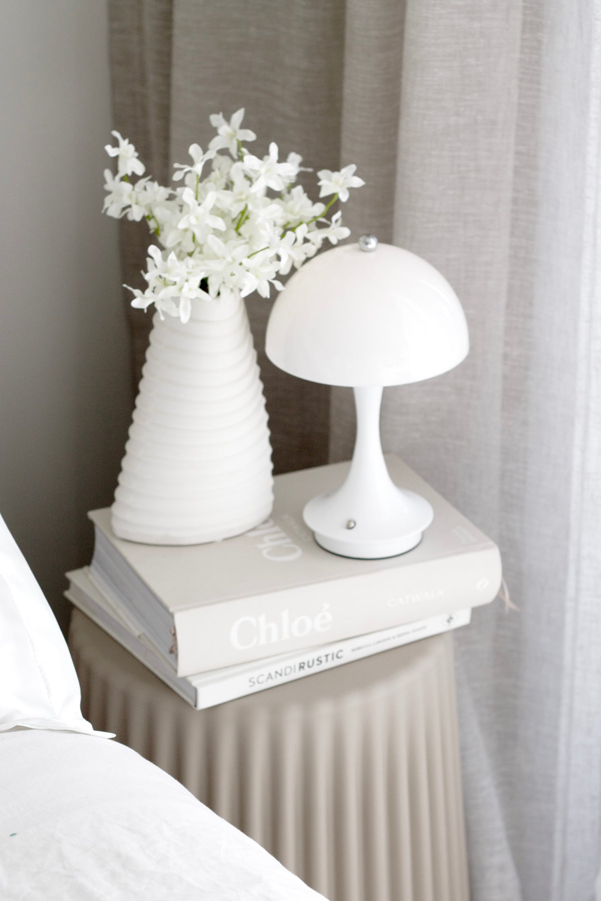 White mushroom lamp unlit on books beside a ribbed vase of white blossom