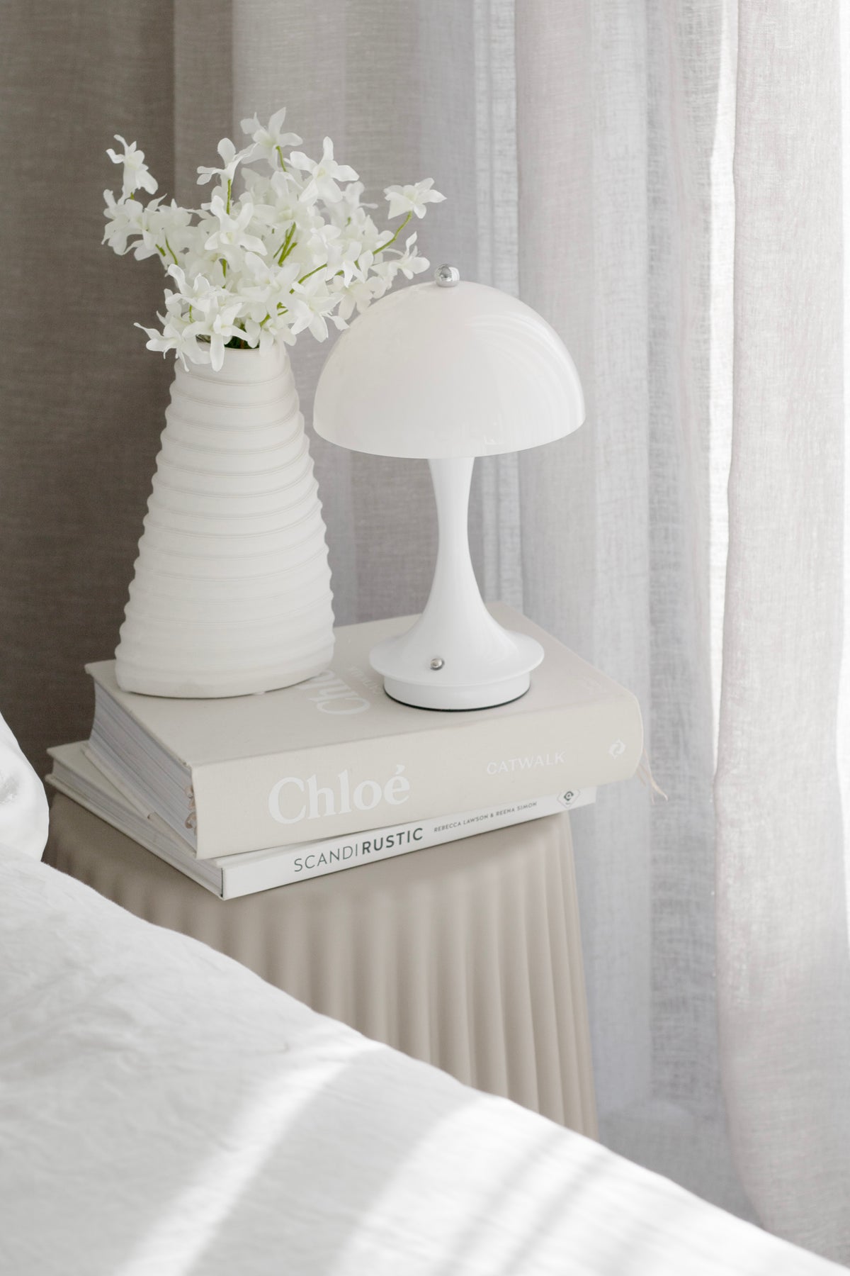 Nude ribbed stool as a bedside table with a ribbed vase of white flowers and a mushroom lamp on books