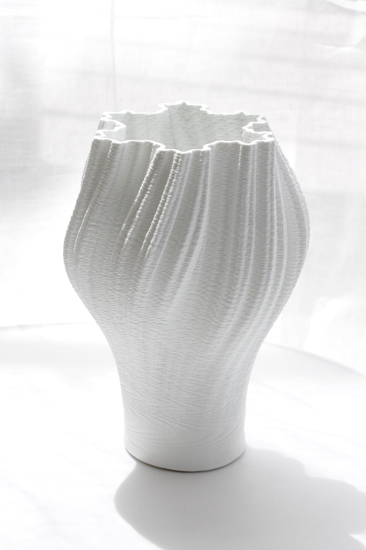 Pleated porcelain vase standing on a pale surface in full sunlight, shadow falling to one side