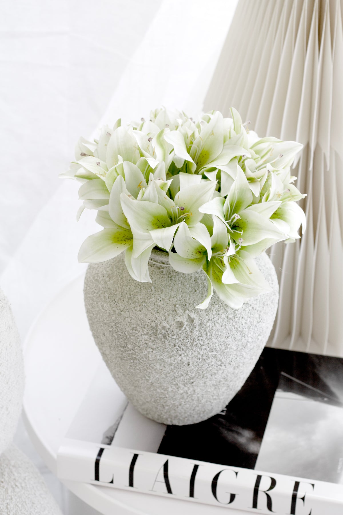 Cream lilies in a speckled stone vase on a book with a pleated paper lamp behind
