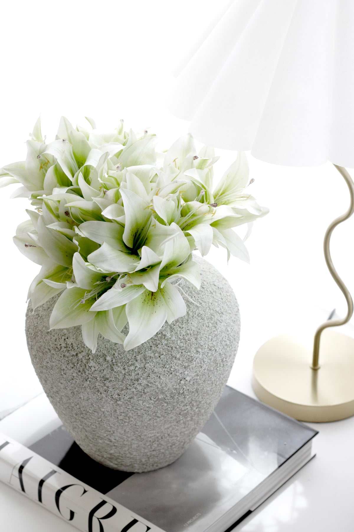 Cream lilies massed in a speckled stone vase beside a brass lamp and stacked books